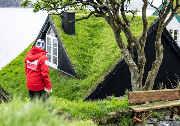 Ein Kreuzfahrer im roten Parka blickt auf eines der grasbedeckten Häuser auf den Färöer Inseln